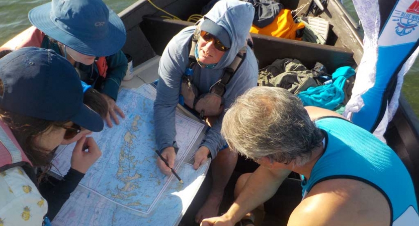 A group of people sitting in a sailboat examine a map that is spread out between them. 
